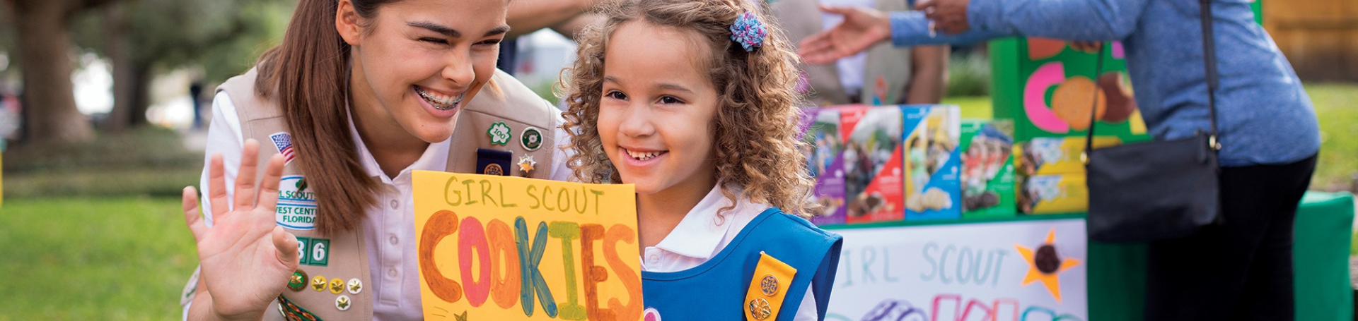  girl scouts selling cookies with one girl in front of booth holding sign that says "girl scout cookie proceeds stay local" 